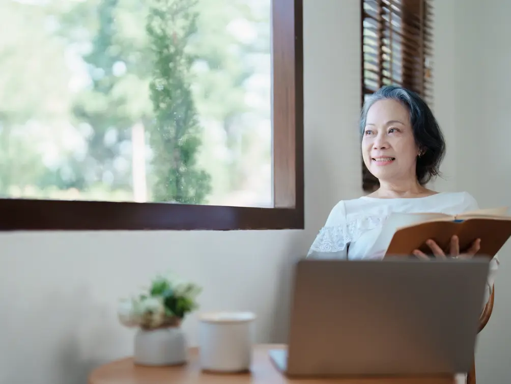 一位亞洲年長女性坐在窗邊，手裡拿著一本書，面帶微笑地望向窗外，桌上有一台筆記型電腦和一盆植物。