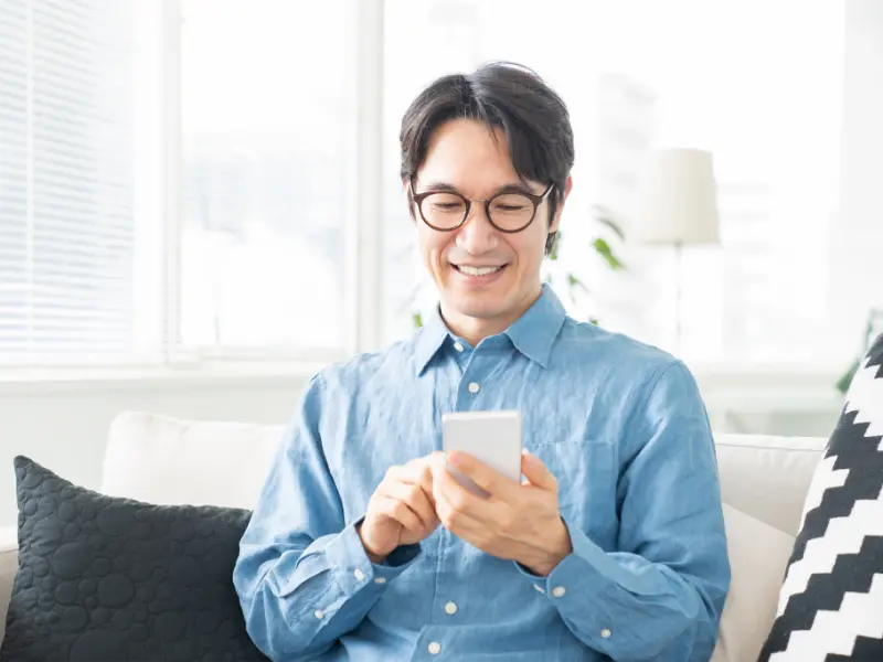 一位戴著眼鏡的男性，身穿藍色襯衫，坐在客廳的白色沙發上，面帶微笑地專注於操作手中的智慧型手機。