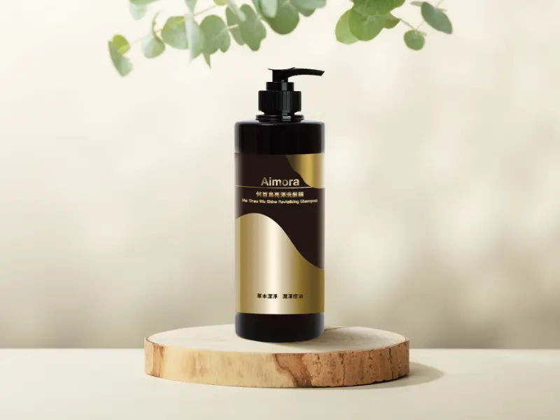 一瓶 Aimora 何首烏煥彩滋養洗髮露，瓶身為黑色，標籤為金屬金色和黑色，放在一片圓形木頭上。背景模糊，有綠色葉子。